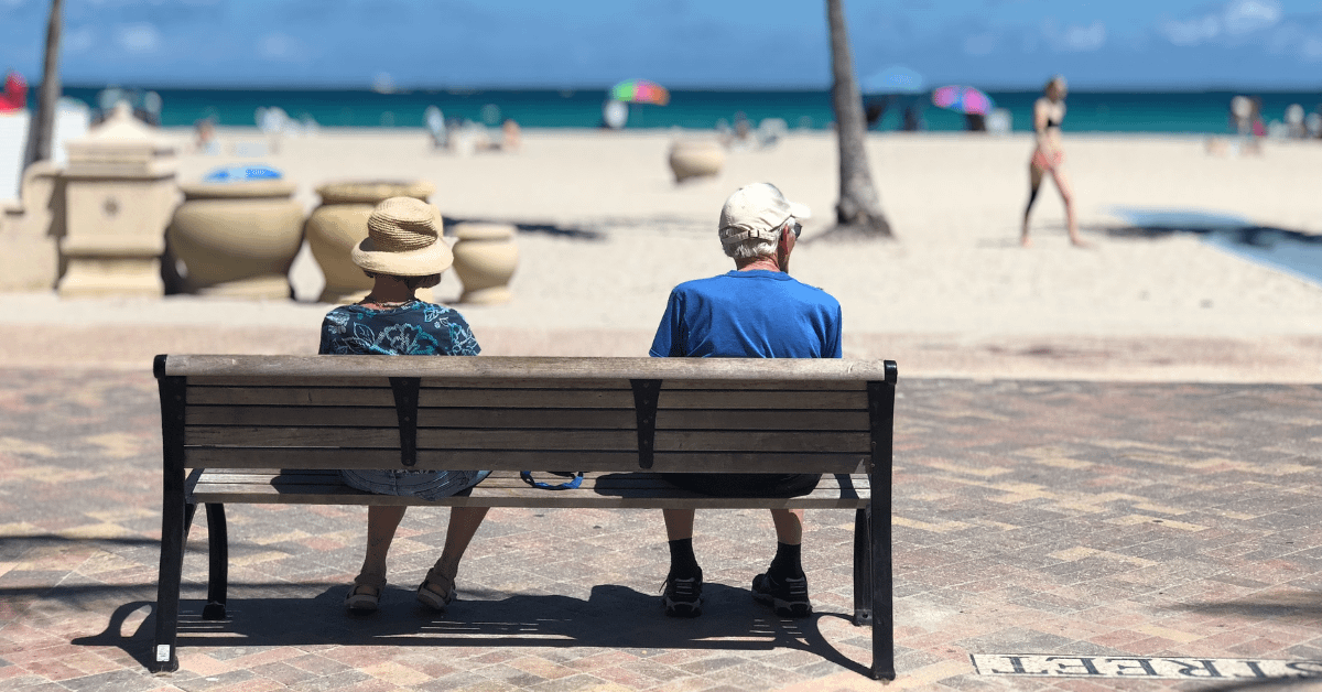 alt="two elderly individuals sitting on a bench overlooking a busy beach"