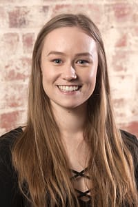 alt="Portrait of Kelly Conlon, smiling against an exposed brick background"